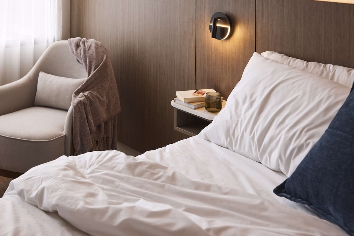 A bedroom with a bed, a chair, a bedside table, and a wall-mounted light fixture.