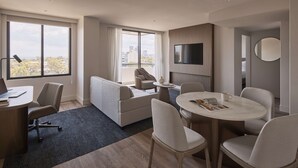 A modern hotel room with a large window, a sofa, a dining table with chairs, a TV mounted on the wall, and a round mirror. - Rydges North Sydney (North Sydney)