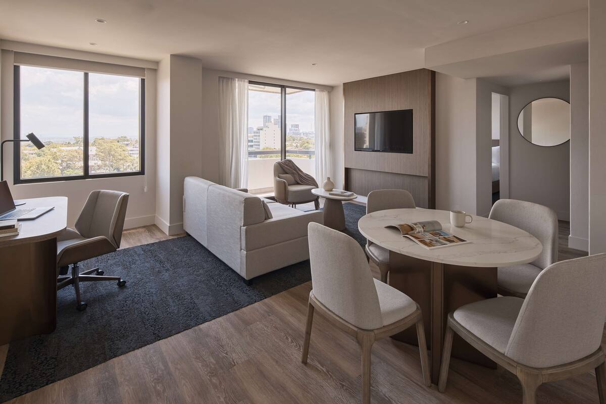 A modern hotel room with a large window, a sofa, a dining table with chairs, a TV mounted on the wall, and a round mirror.