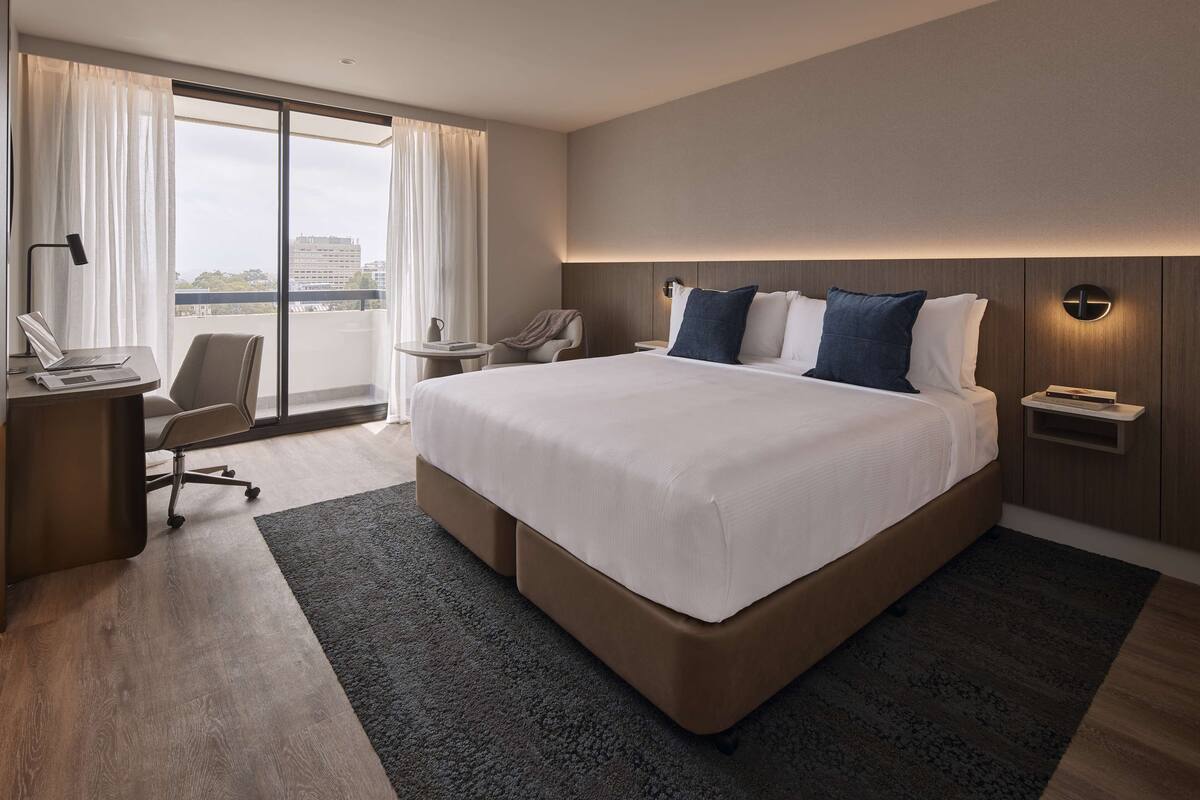 A modern hotel room with a large bed, a desk with a laptop, a chair, and a view of the city through large windows.