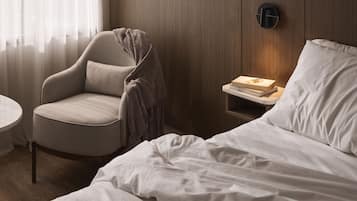 Une chambre d’hôtel avec un lit, une chaise, une table de chevet avec des livres et un luminaire fixé au mur.