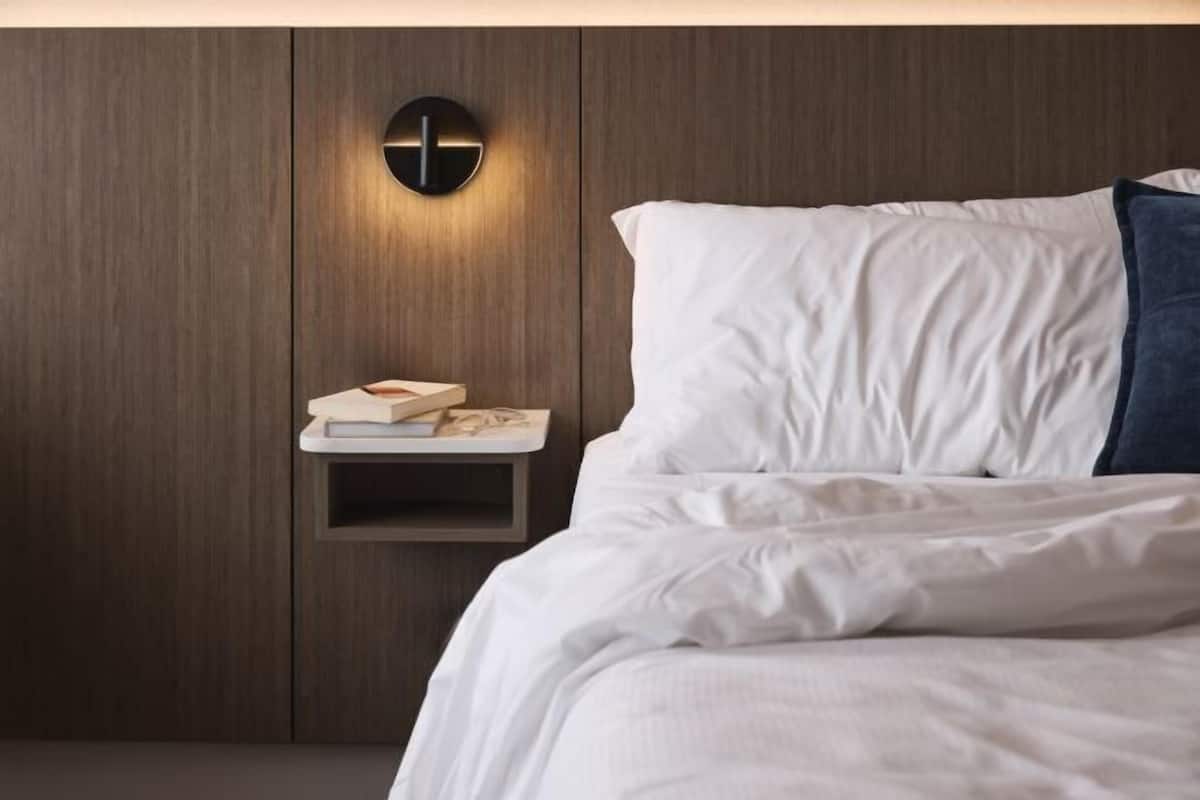 A bedroom with a bed, white bedding, a nightstand with a lamp, and a small shelf with books.