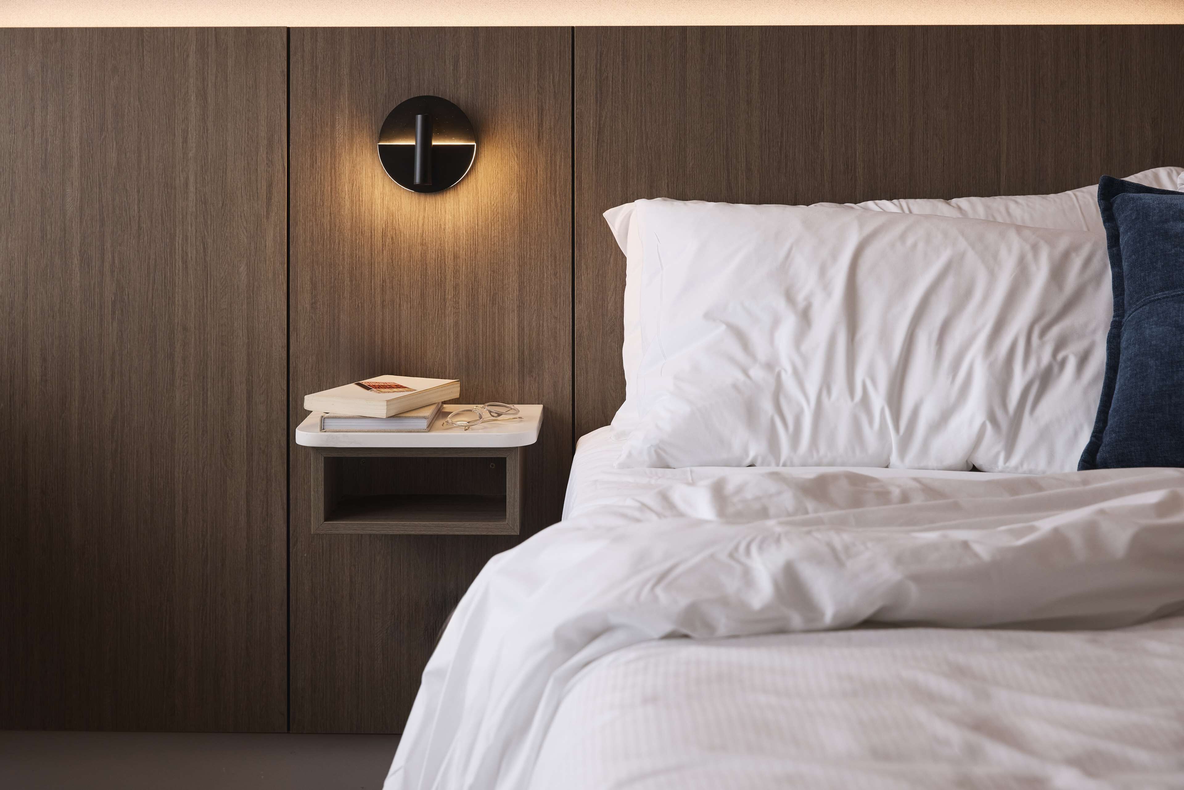 A neatly made bed with white bedding and a blue pillow, a bedside table with books and glasses, and a wall-mounted light fixture.