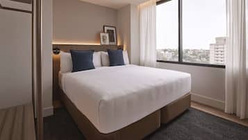 A hotel room with a large bed, a view of the city through a window, and a door to the left.