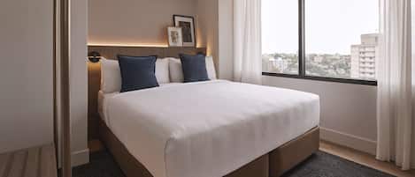 A hotel room with a large bed, a view of the city through a window, and a door to the left.