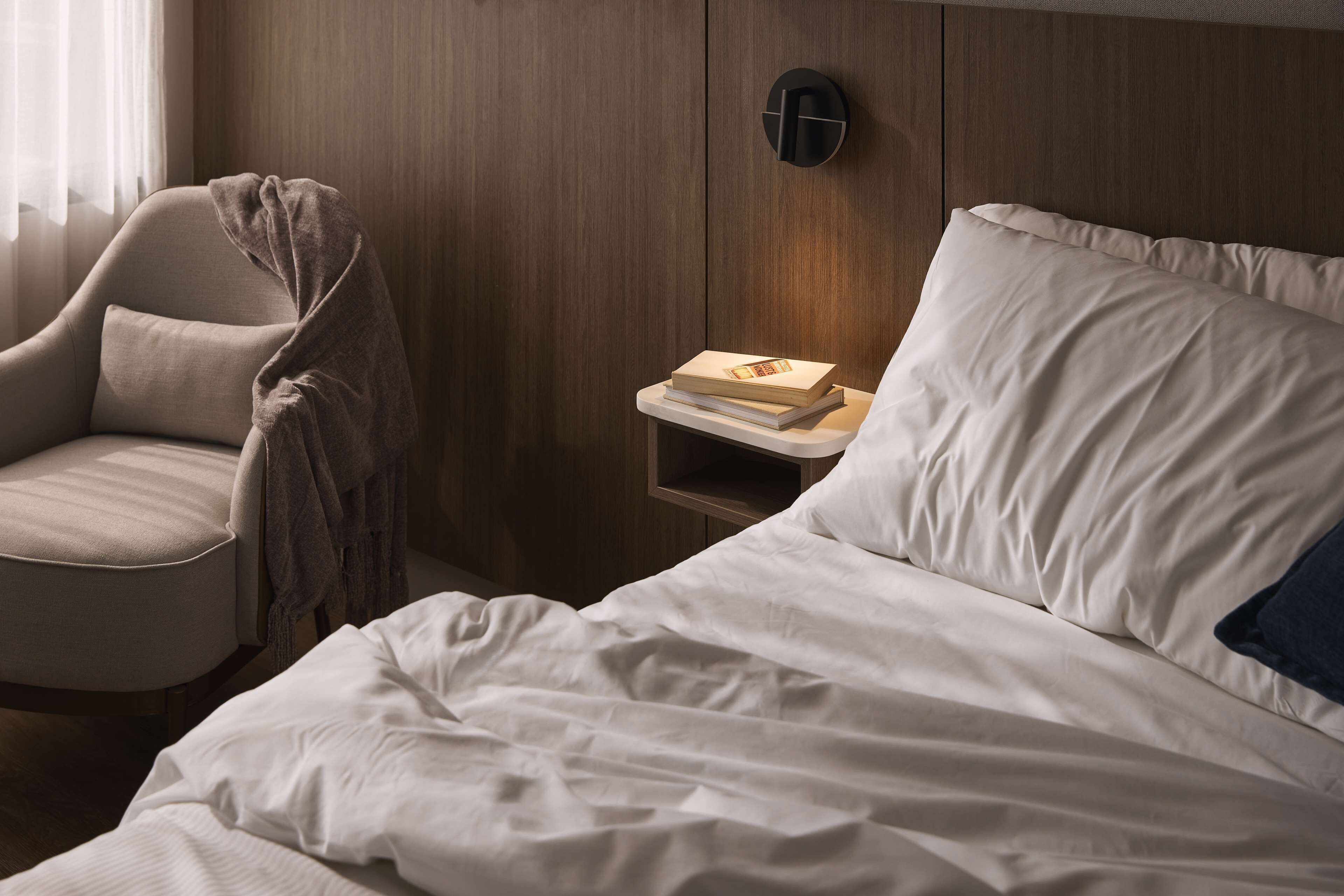 A neatly made bed with white linens, a bedside table with a book, and a chair with a blanket.