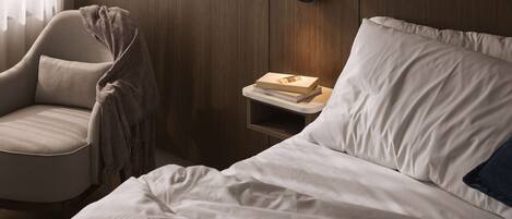 A neatly made bed with white linens, a bedside table with a book, and a chair with a blanket.