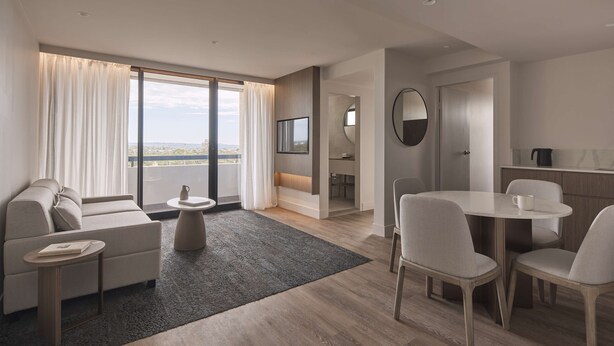 A modern hotel room with a sofa, a dining table with chairs, a small coffee table, a large window with curtains, a TV, and a bathroom visible through an open door. - Rydges North Sydney (North Sydney)