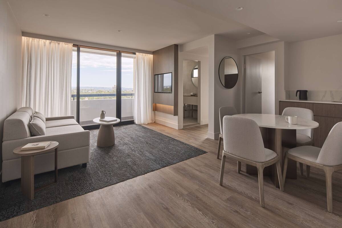 A modern hotel room with a sofa, a dining table with chairs, a small coffee table, a large window with curtains, a TV, and a bathroom visible through an open door.