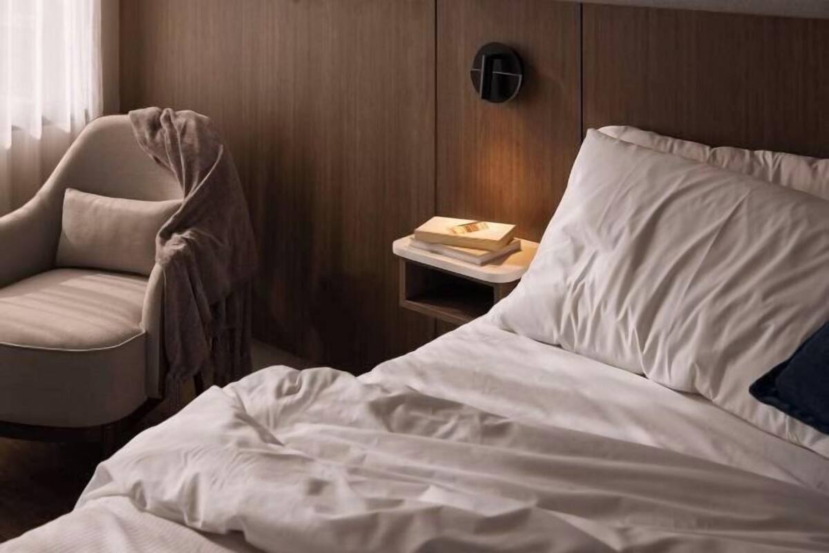 A hotel room with a bed, a chair, a nightstand with a book, and a wall-mounted light.