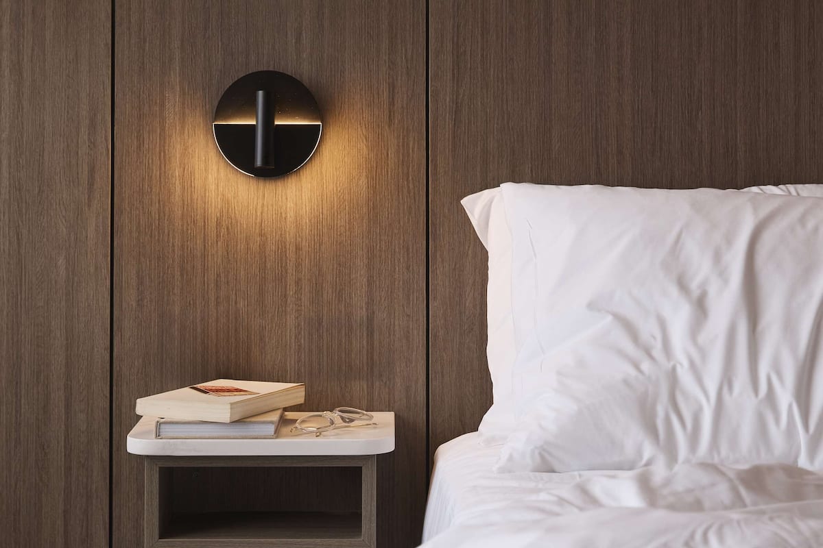 A bedroom with a bed, white bedding, a nightstand with a book and glasses, and a wall-mounted light fixture.