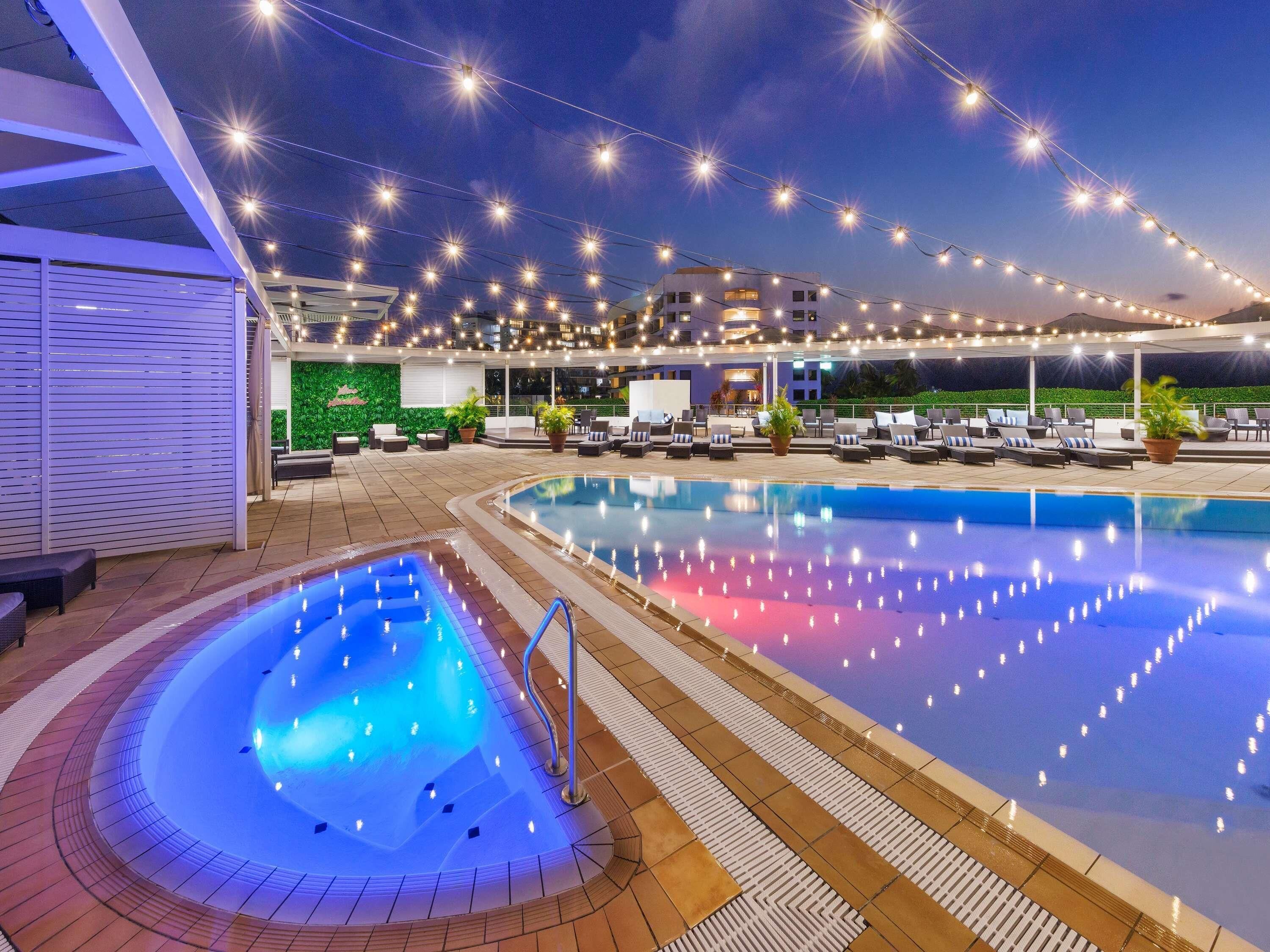 Uma área de piscina com duas piscinas, cadeiras de sol e luzes decorativas.