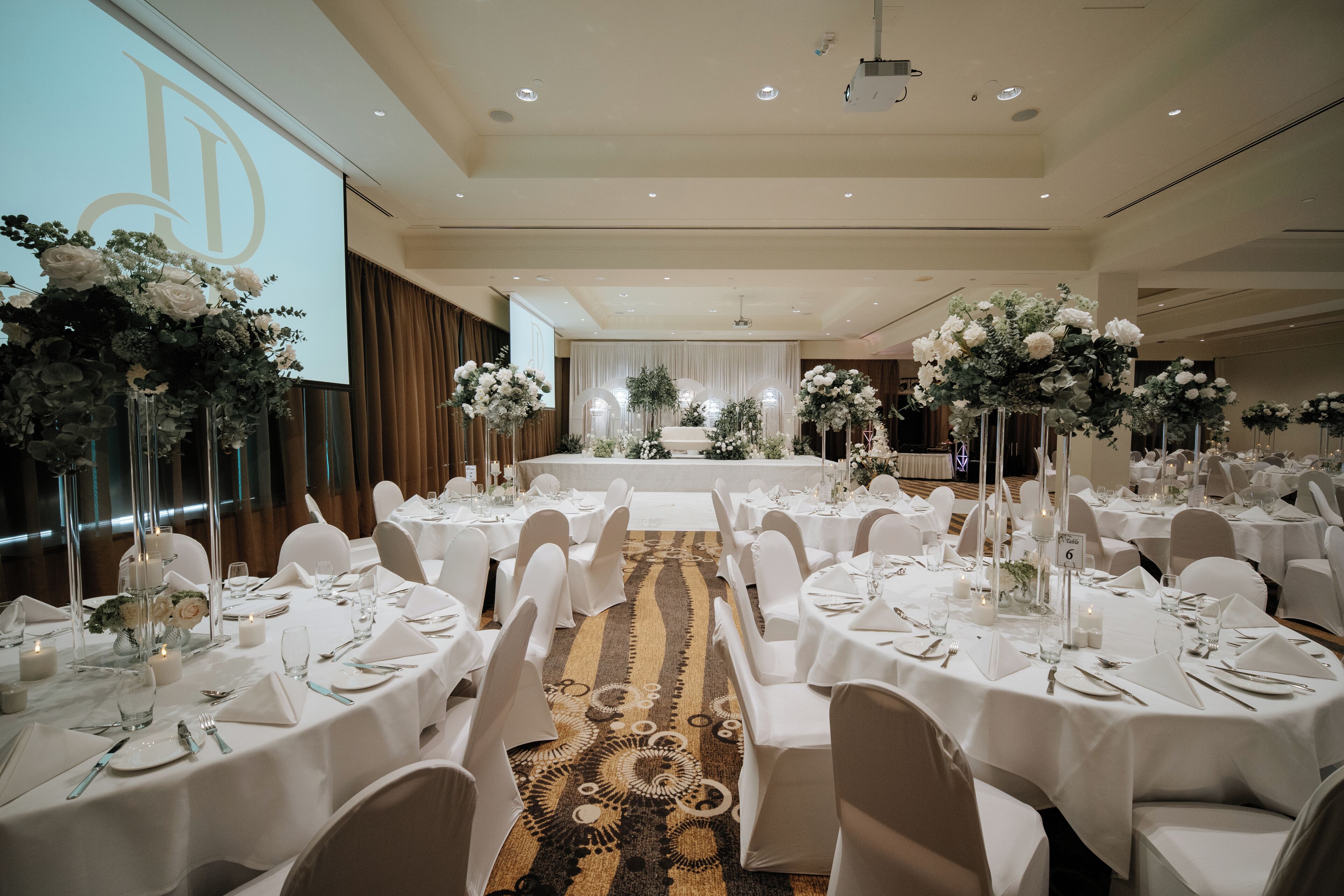 Un salón de banquetes con mesas redondas dispuestas para un evento formal, con manteles blancos, centros de mesa florales y suelo alfombrado.