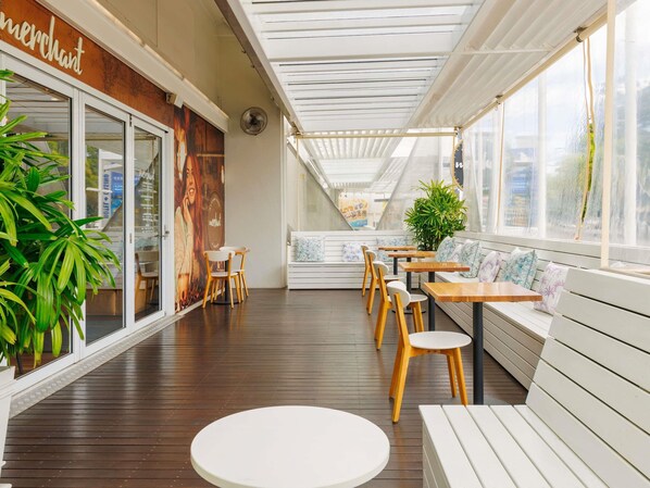 A covered outdoor seating area with wooden tables and chairs, a white bench, and a large potted plant.