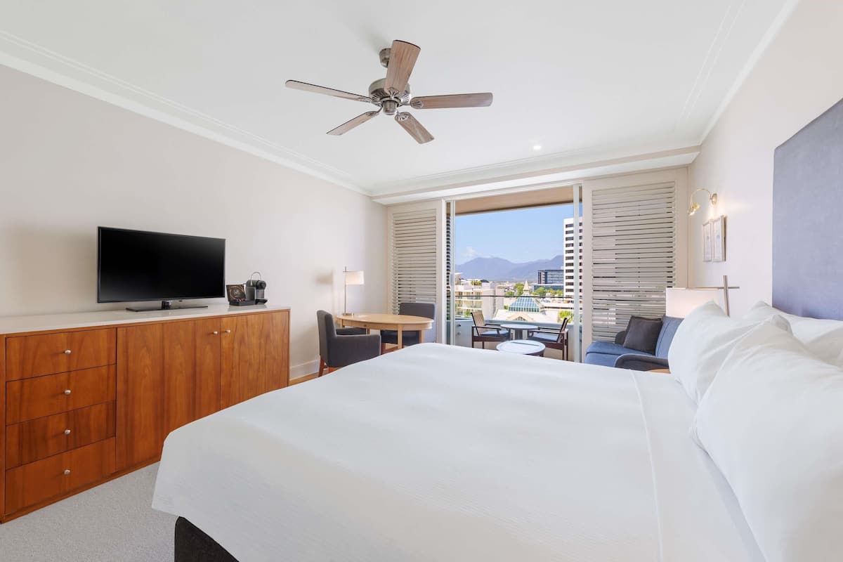 Hotelkamer met een bed, televisie, bureau met stoel, balkon met uitzicht en plafondventilator.