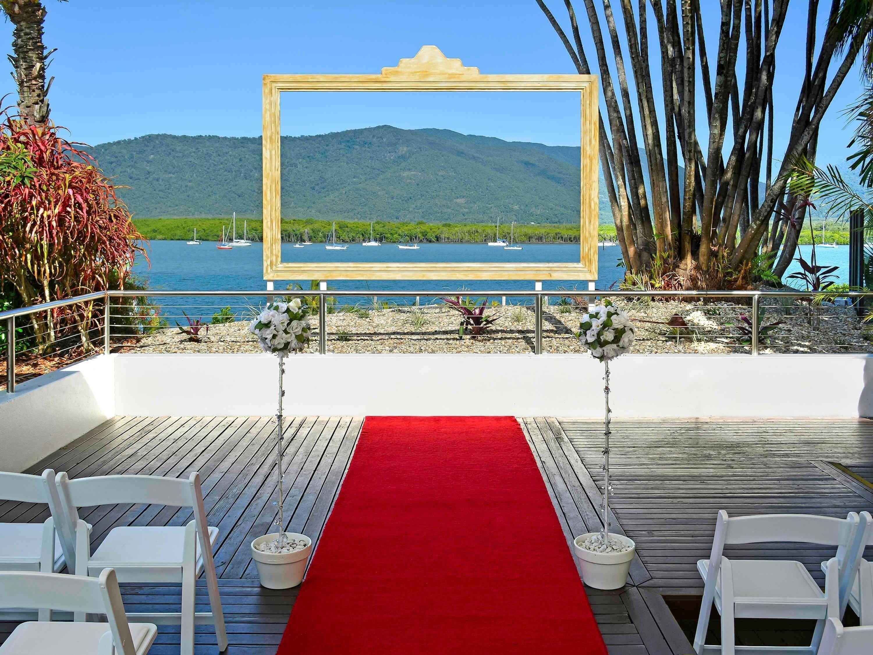 An outdoor wedding setup with a red carpet, white chairs, and a wooden frame with a view of boats and mountains.