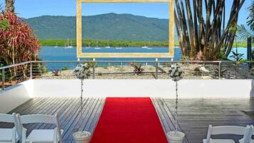 An outdoor wedding setup with a red carpet, white chairs, and a wooden frame with a view of boats and mountains.