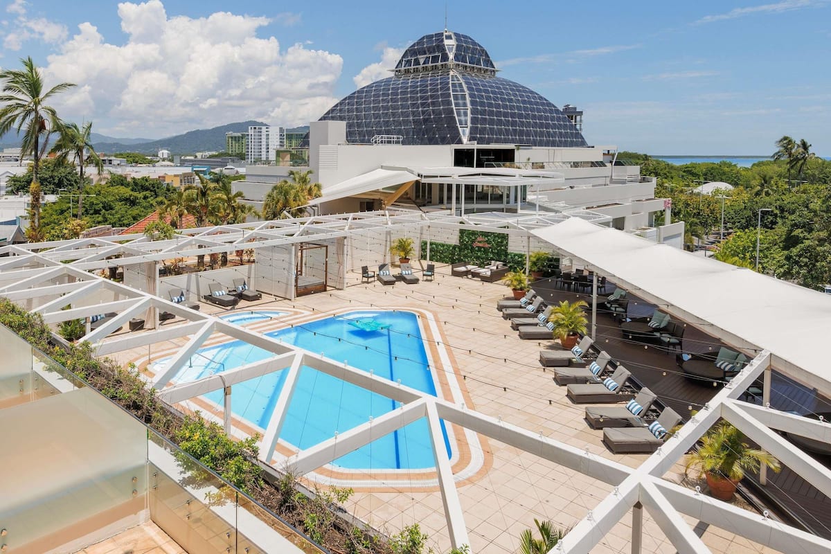 A rooftop pool area with lounge chairs, a swimming pool, and a large building with a dome-like structure in the background.