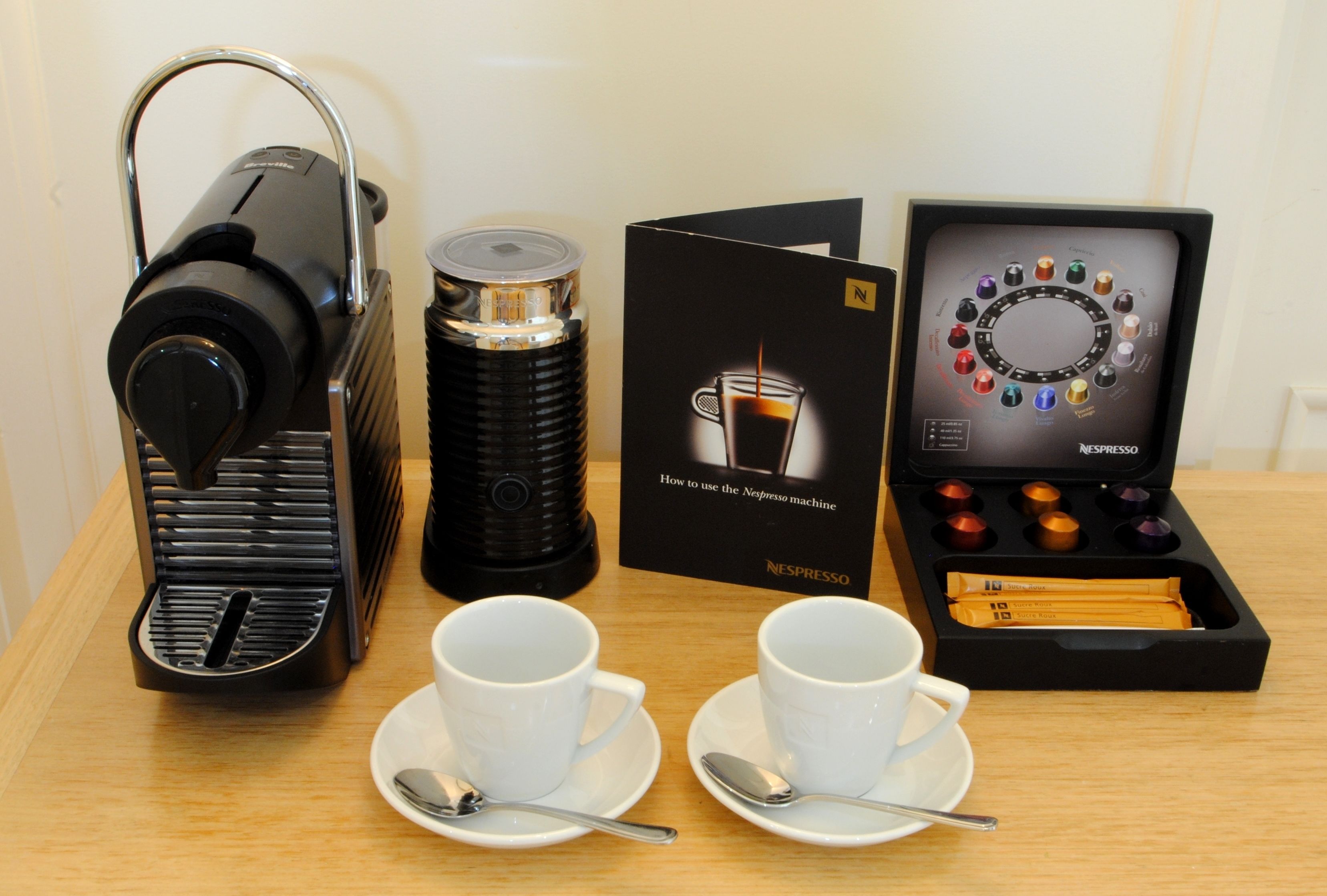 En kaffemaskin, två koppar med teskedar, en förpackning med kaffekapslar och en bruksanvisning.