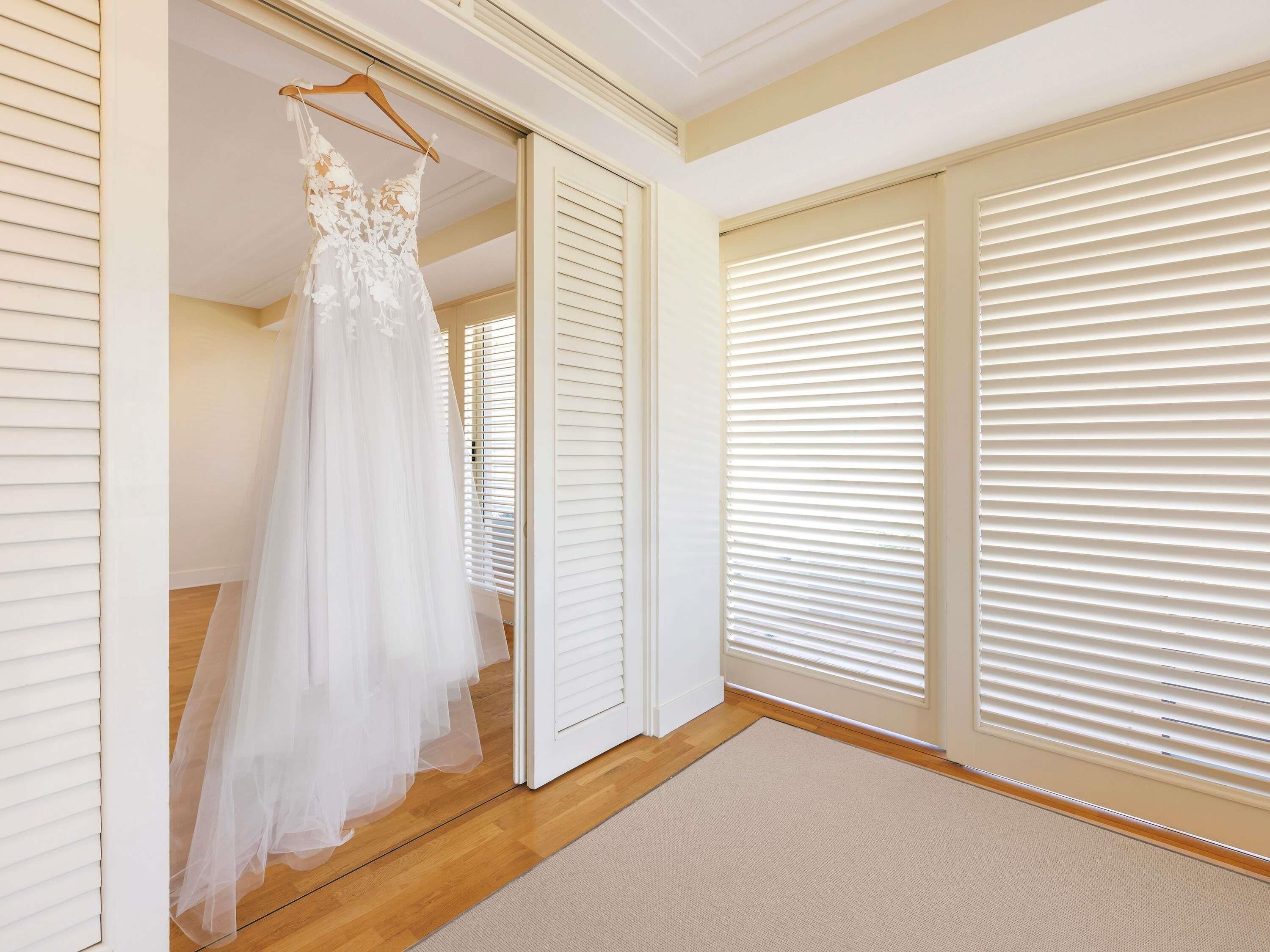 A bridal gown hanging in a room with wooden floors, white walls, and large windows with blinds.