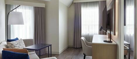 A hotel room with a sofa, a desk, a chair, a lamp, and a TV.
