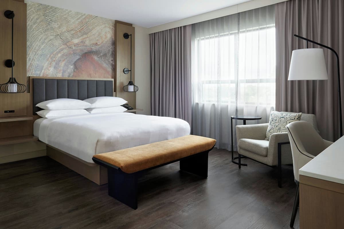 Habitación de hotel moderna con una cama grande, un banco, una silla, un escritorio y un ventanal con cortinas.