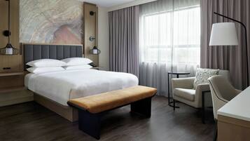 A modern hotel room with a large bed, a bench, a chair, a desk, and a large window with curtains.