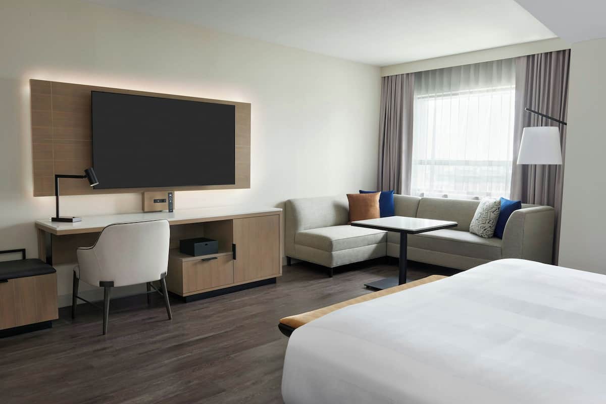Una habitación de hotel moderna con una cama grande, un escritorio con silla, un sofá con cojines, un televisor de pantalla plana montado en la pared y una ventana con cortinas.