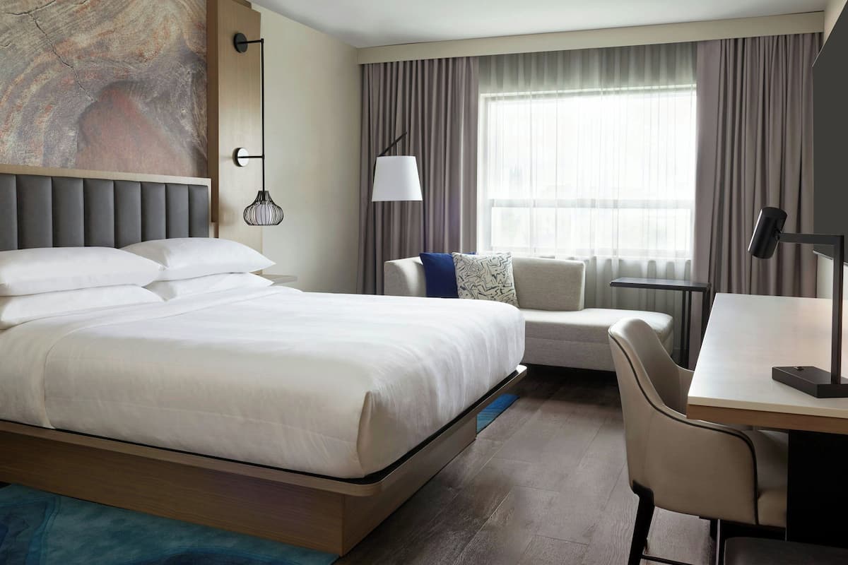 Habitación de hotel con una cama grande, un escritorio, una silla, una lámpara y vistas al exterior a través del ventanal.