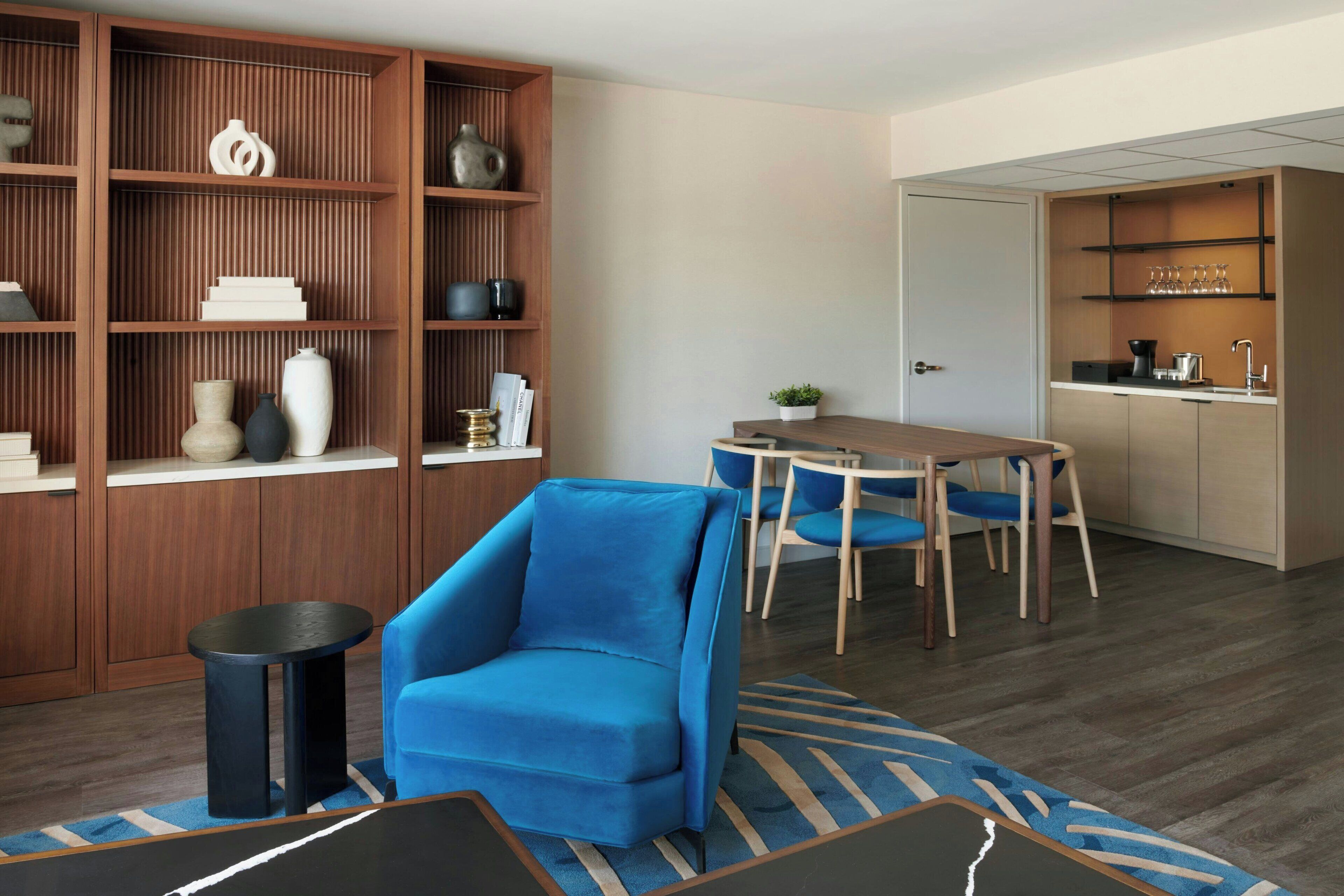 Una sala de estar moderna con un sillón azul, una estantería de madera, una zona de comedor con mesa y sillas, y una cocina con lavaplatos y encimera.