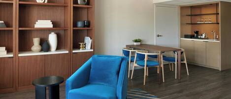 Una sala de estar moderna con un sillón azul, una estantería de madera, una zona de comedor con mesa y sillas, y una cocina con lavaplatos y encimera.