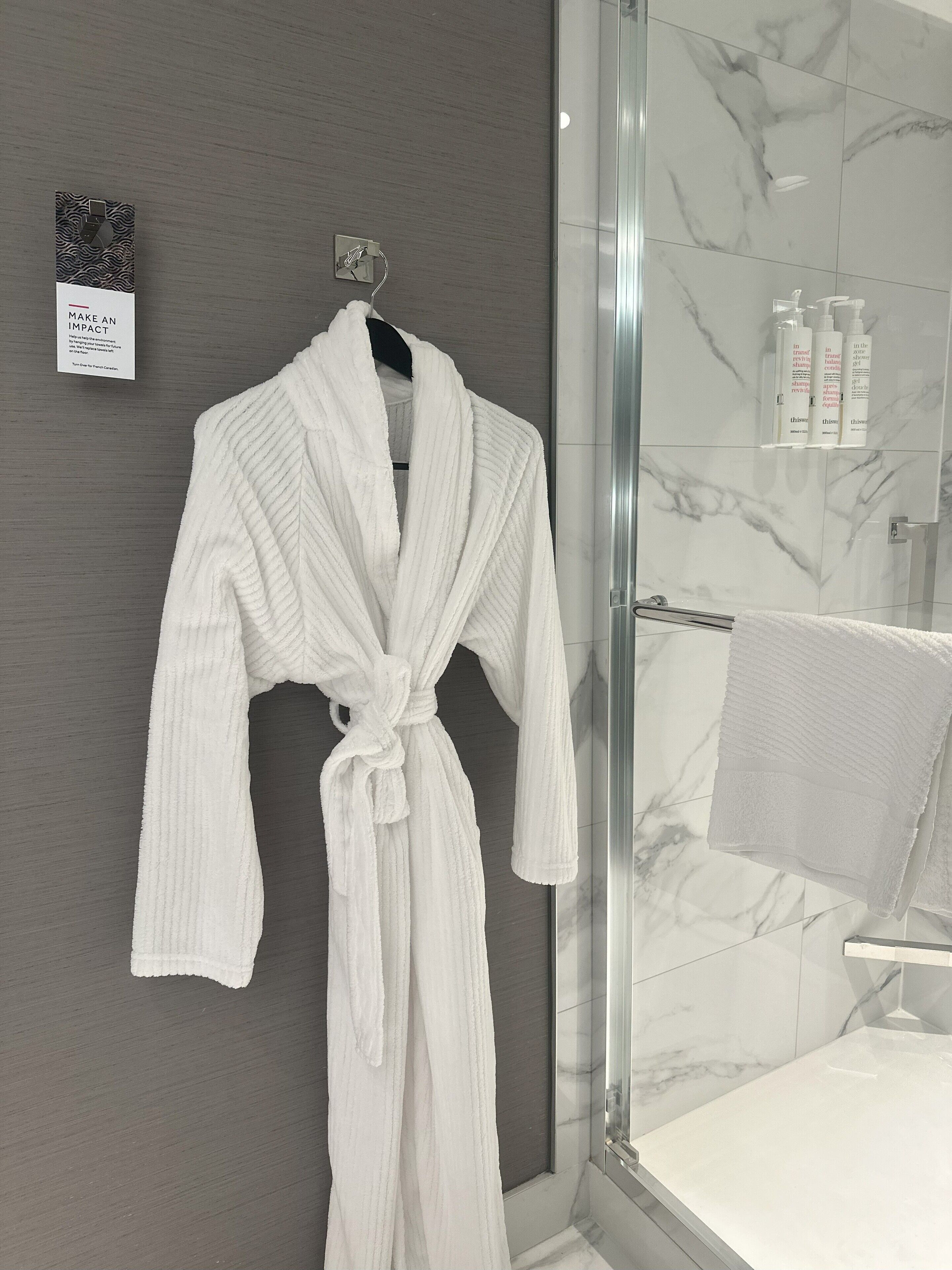 A white bathrobe hanging on a hook in a bathroom with a glass shower enclosure and marble tiles.