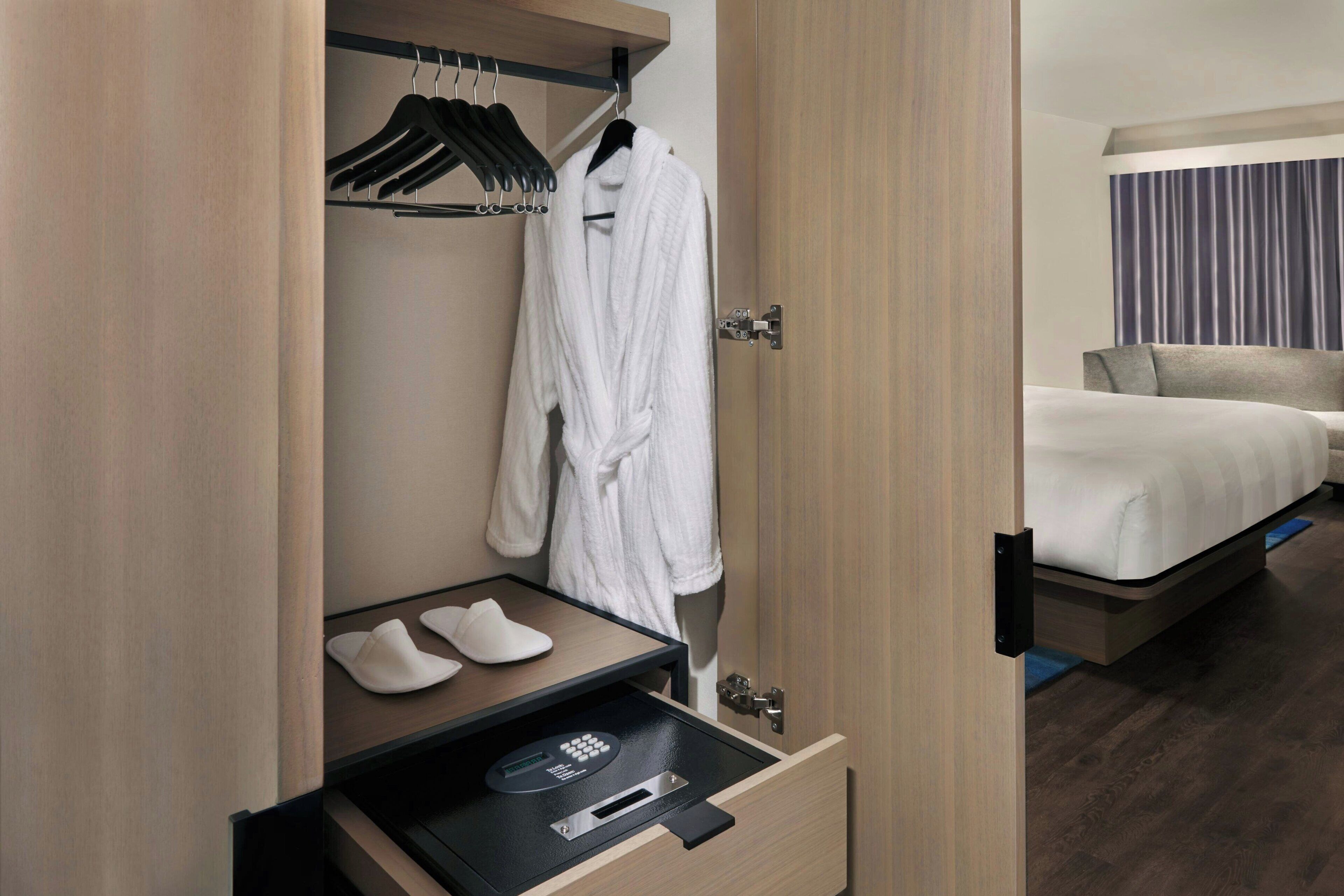 Quarto de hotel com guarda-roupa de madeira, um roupão pendurado no interior, um par de chinelos numa prateleira e uma cama com cabeceira cinzenta.