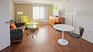 A hotel room with a wooden floor, a sofa, a yellow armchair, a small round table, a white dining table, and a television on a cabinet. - Holiday Inn Express Indianapolis-Fishers by IHG (Fishers)