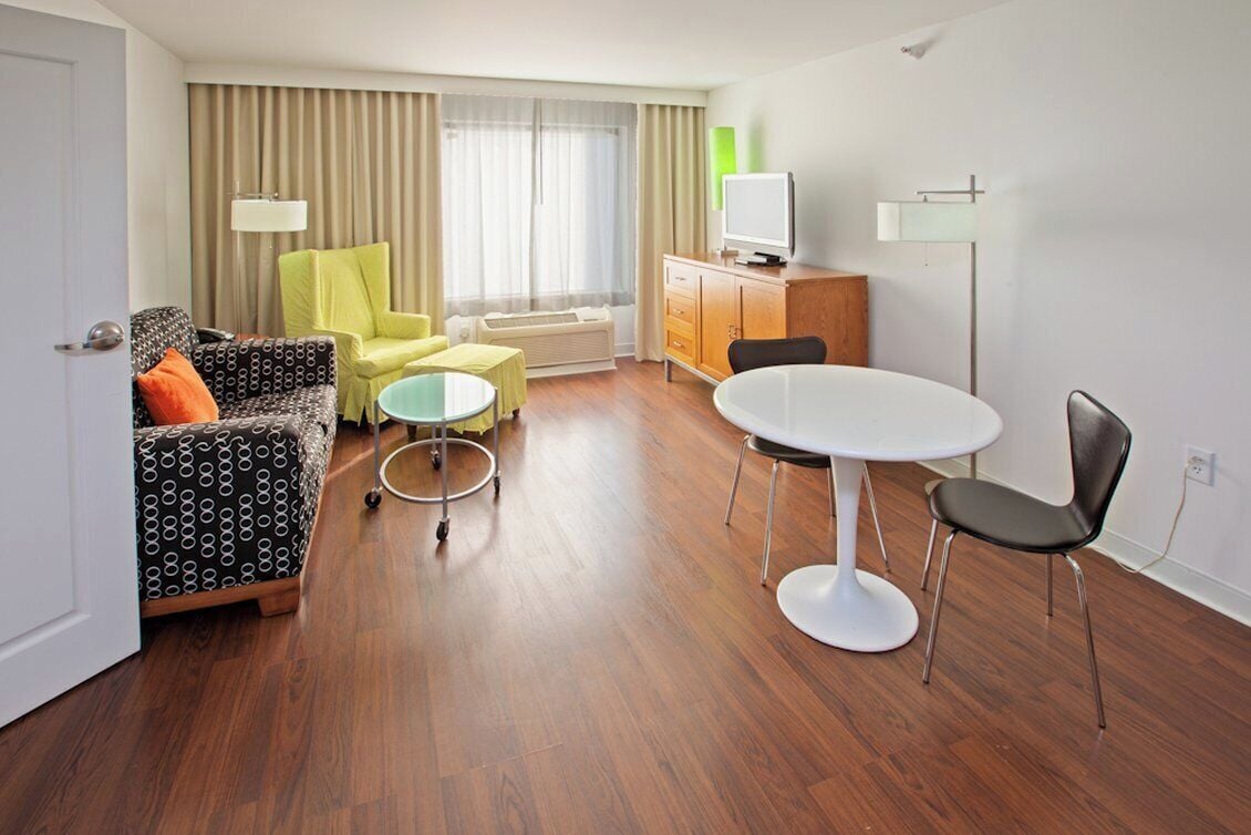 A hotel room with a sofa, a yellow armchair, a small round table, a white dining table, and a TV on a cabinet.