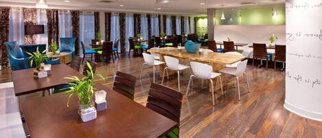 Un comedor moderno con varias mesas y sillas, una gran mesa de madera en el centro, y plantas decorativas.
