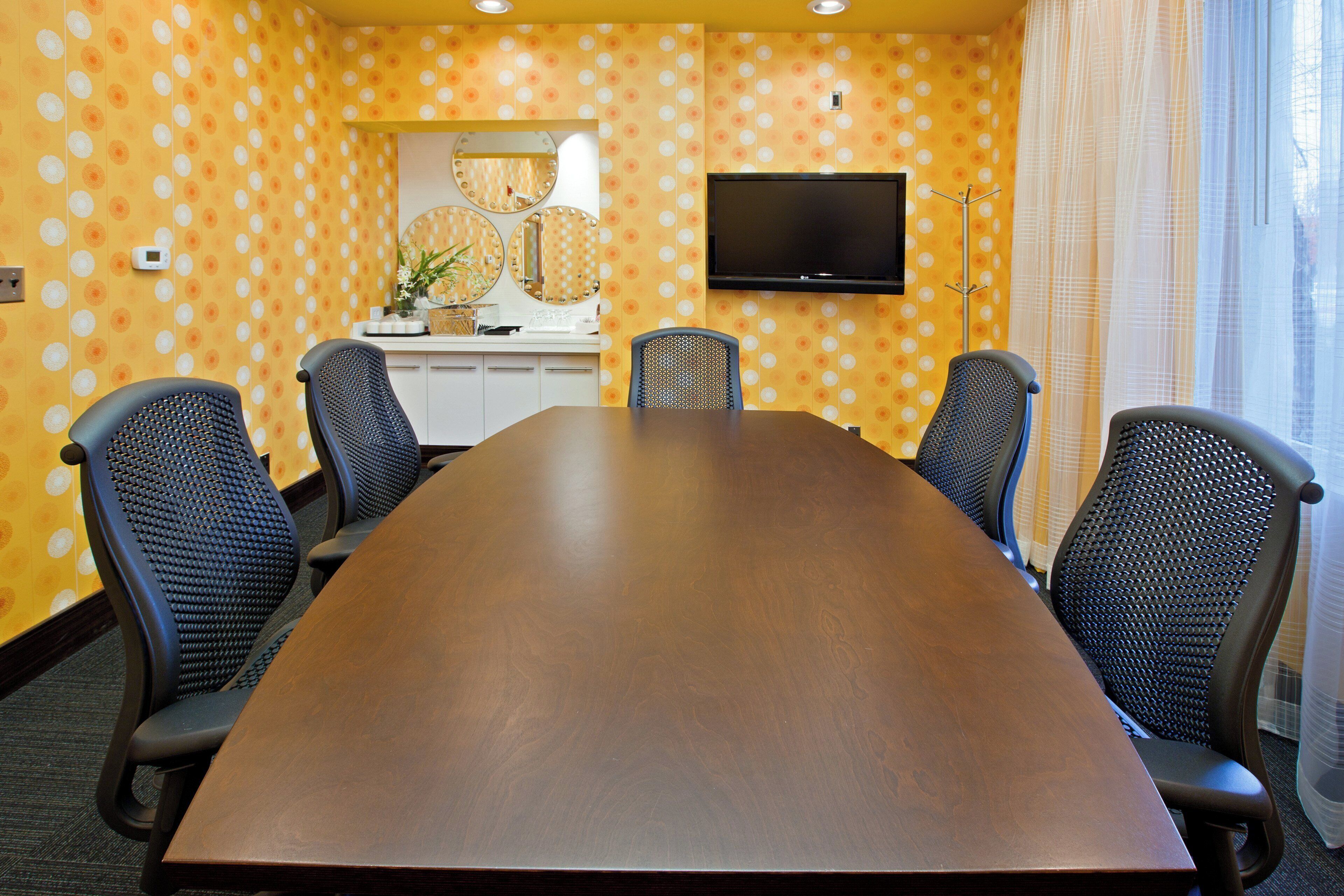 Een vergaderruimte met een lange tafel, zes stoelen, een televisie en een spiegel aan de muur.