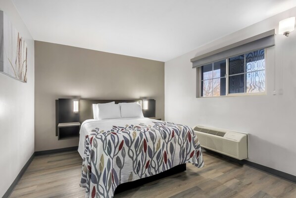 A bedroom with a bed, bedside tables, a window with patterned blinds, and a wall-mounted air conditioning unit. - Red Roof Inn Globe - Miami, AZ (Miami)