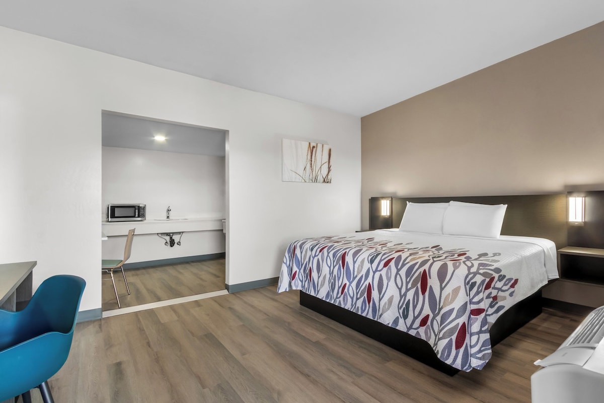 A modern hotel room with a wooden floor, a bed with a patterned bedspread, a desk with a chair, a microwave, and a small kitchenette.