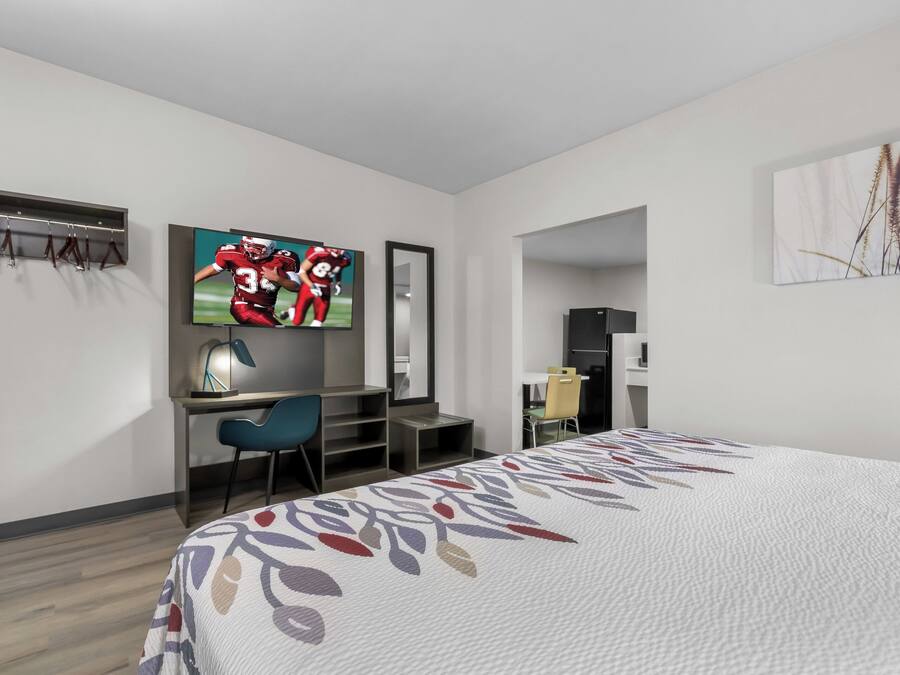 Ett modernt hotellrum med en säng, ett skrivbord med en stol, en tv som är monterad på väggen och ett kylskåp i hörnet.
