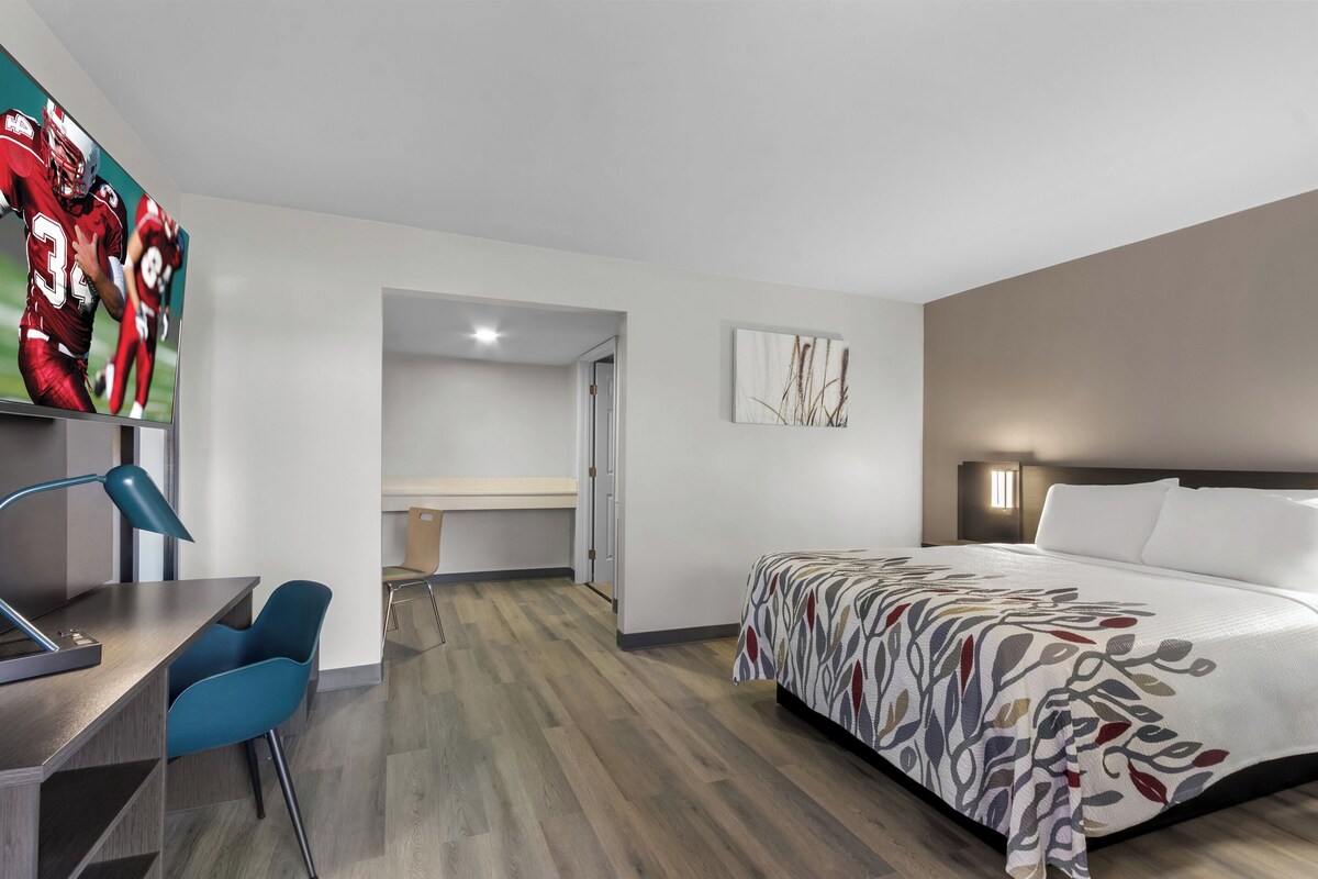 A modern hotel room with a bed, desk, and chair. There is a television on the wall displaying a sports game.