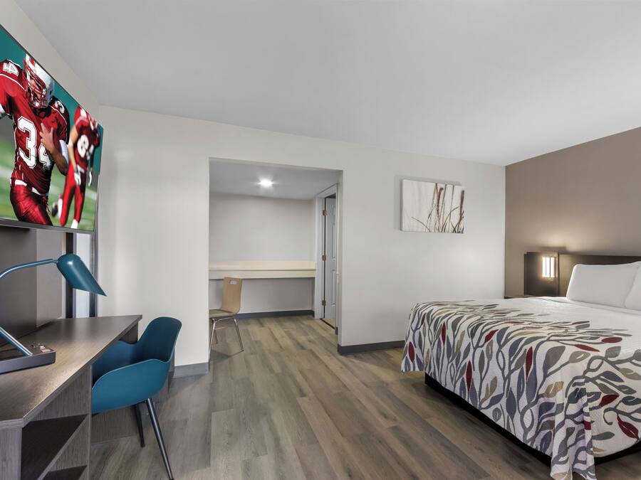 A modern hotel room with a bed, desk, and chair. There is a television on the wall displaying a sports game.