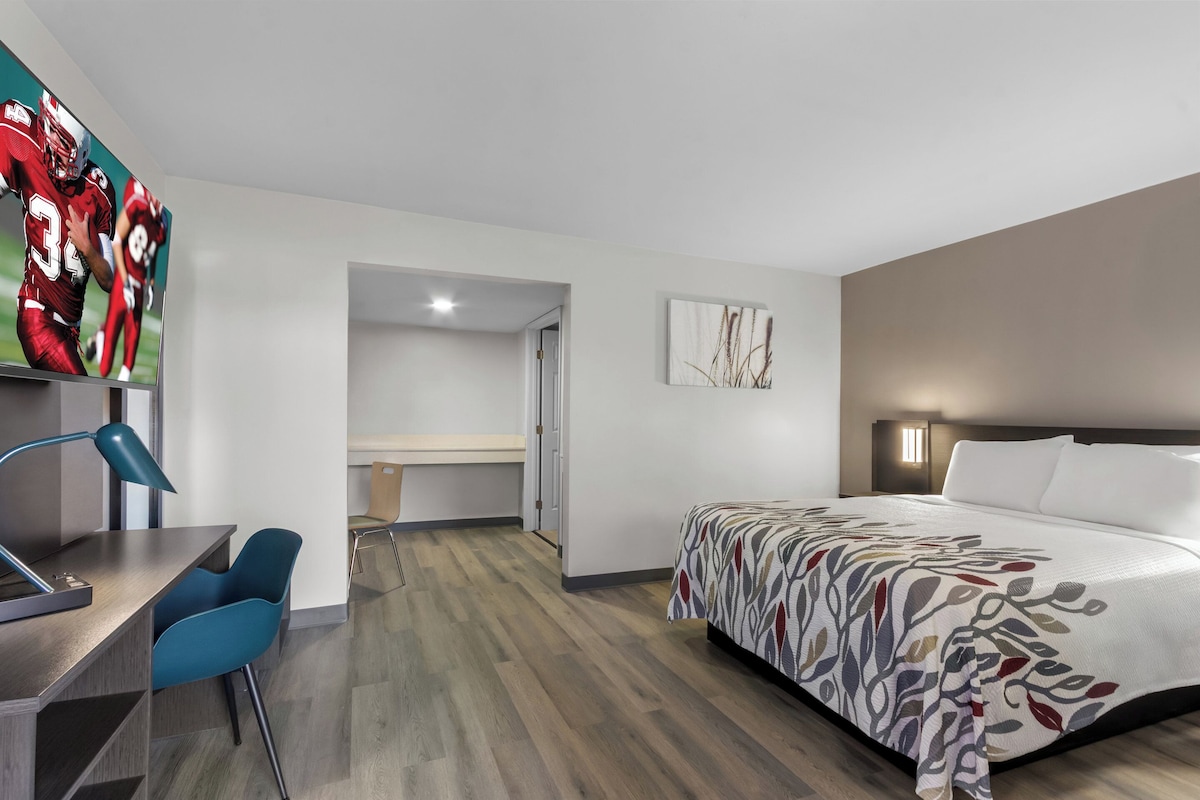 A modern hotel room with a bed, desk, and chair. There is a television on the wall displaying a sports game.