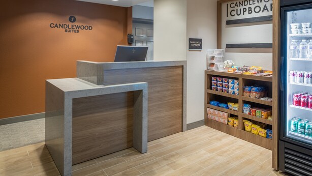 Un comptoir d’accueil avec un ordinateur portable, une enseigne indiquant « CANDLEWOOD SUITES » et un « CANDLEWOOD CUPBOARD » contenant des collations et des boissons.