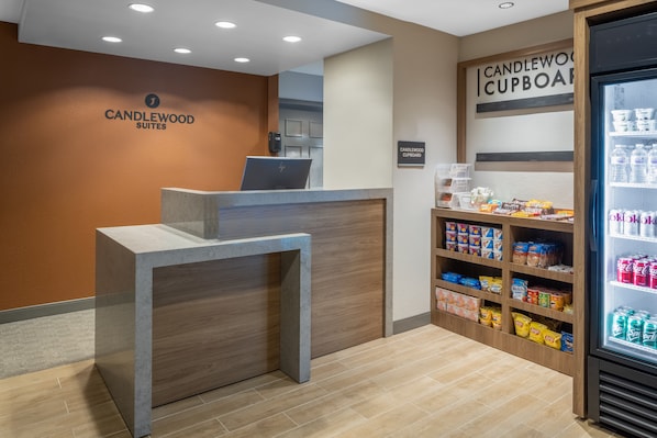 Un mostrador de recepción con un ordenador portátil, un cartel que dice "CANDLEWOOD SUITES" y un "CANDLEWOOD CUPBOARD" con aperitivos y bebidas.