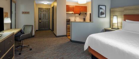 A hotel room with a bed, desk, chair, kitchenette, and a hallway leading to another room.