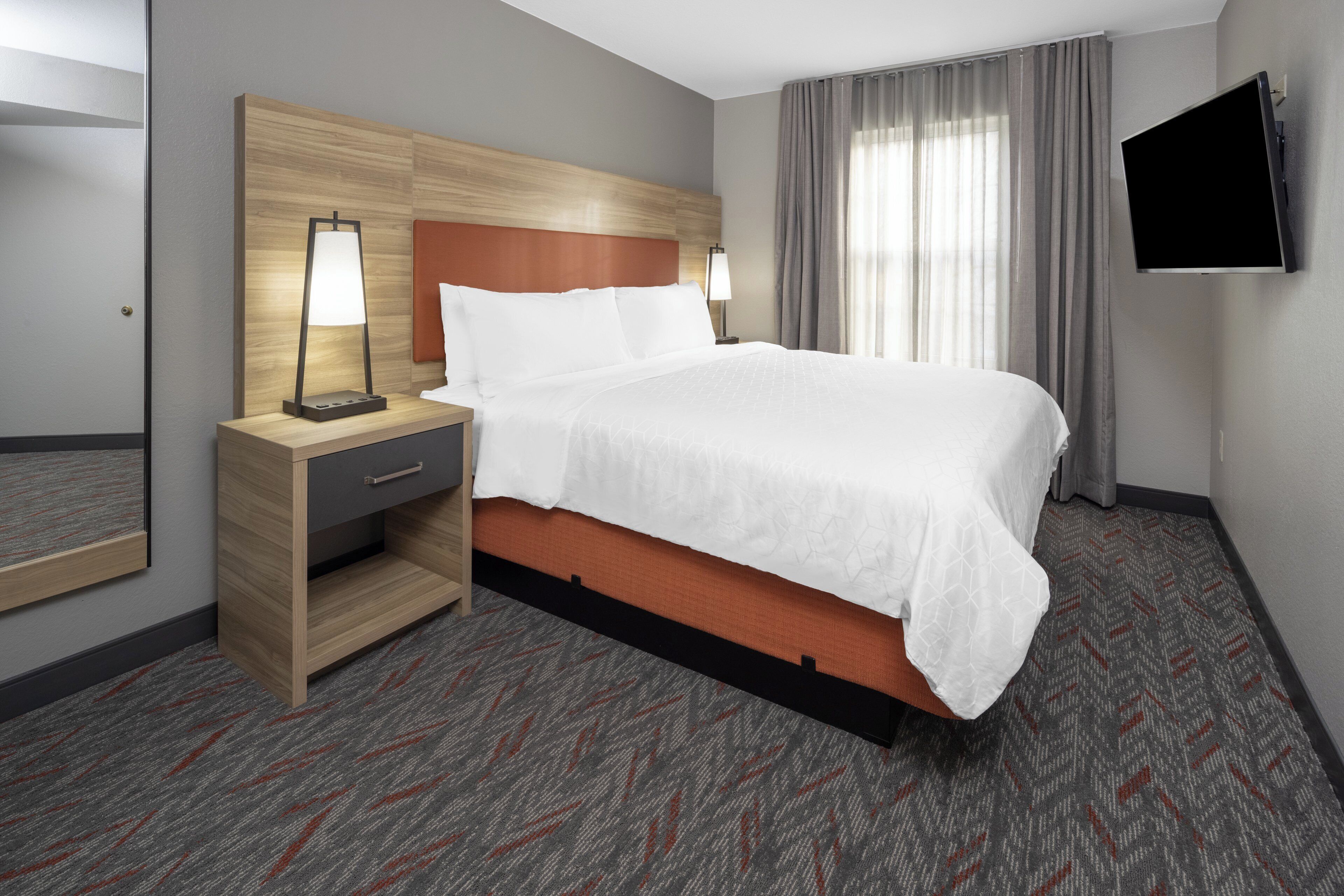 A hotel room with a bed, nightstand, lamp, TV, and a mirror.