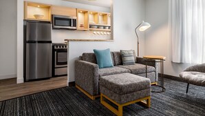 Een moderne woonkamer met een grijze bank, een voetenbankje en een kleine tafel. Er is een keuken met een koelkast, magnetron en oven. Een vloerlamp en een raam met gordijnen zijn ook zichtbaar.