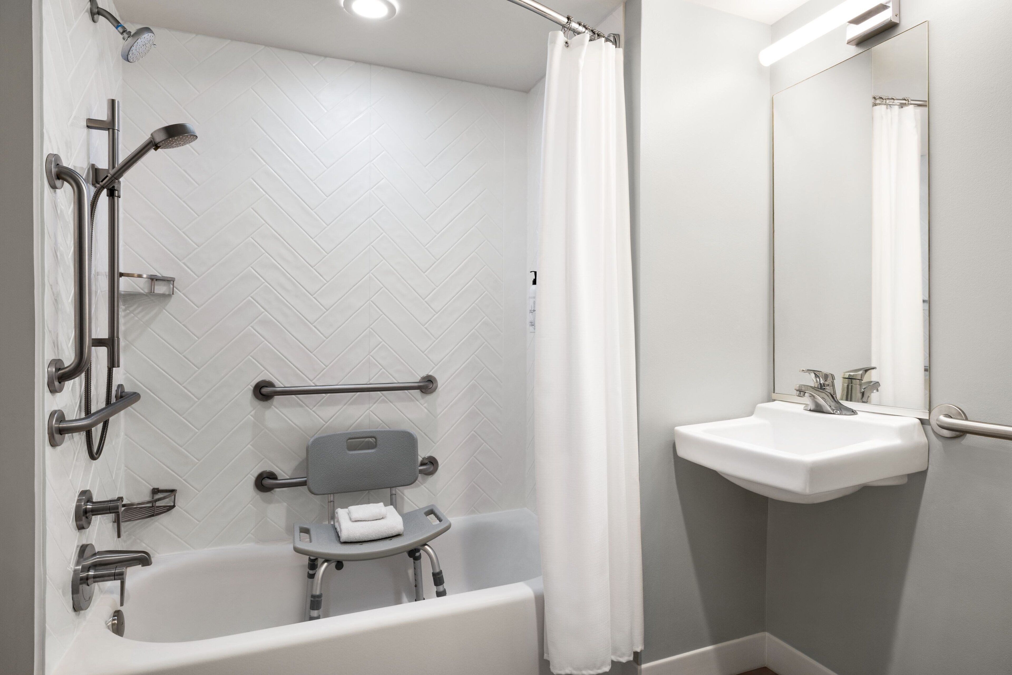 A modern bathroom with a white herringbone tile wall, a white bathtub with grab bars, a white sink with a mirror, and a shower with a white curtain.