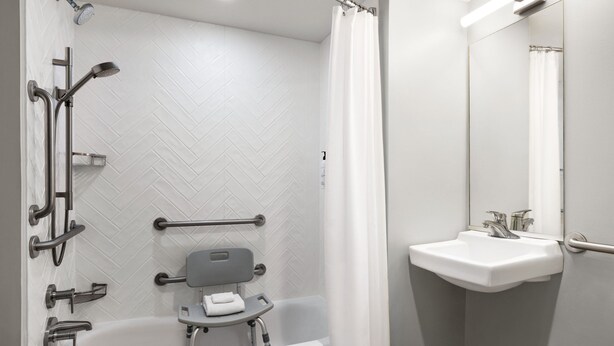 Een moderne badkamer met een witte muur betegeld in een visgraatmotief, een witte badkuip met handgrepen, een witte wastafel met spiegel, en een douche met een wit gordijn.